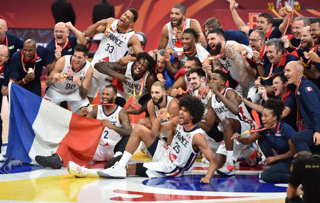 L’équipe nationale masculine de basketball de France célèbre sa médaille de bronze sur le terrain, entourée de joueurs en maillots blancs arborant fièrement le drapeau tricolore. Sourires, accolades et joie collective marquent ce moment de fierté sportive.